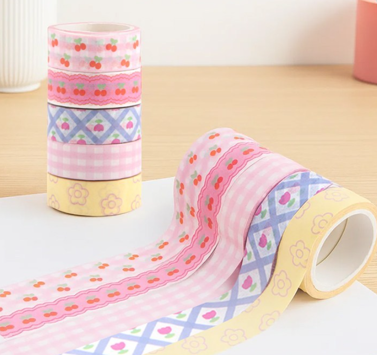 Four rolls of decorative washi tape with pastel patterns, including pink cherry, pink gingham, blue lattice, and yellow floral designs, arranged on a light surface.