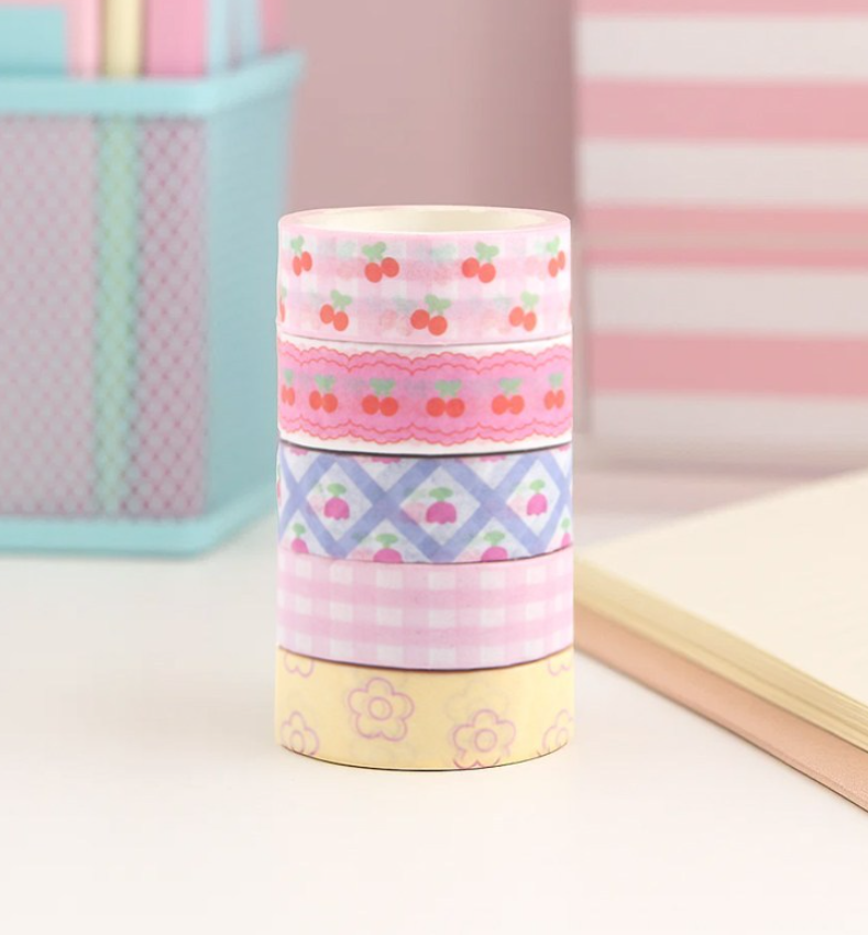 Four rolls of decorative washi tape stacked vertically on a white surface. The tapes feature cherry and gingham patterns in pink and yellow, with one roll showing a lavender and blue diamond pattern.
