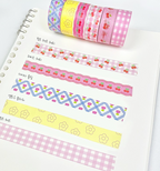 A set of decorative washi tapes with various patterns including cherry prints, gingham, and geometric designs, shown both rolled up and laid out on a white spiral notebook page.