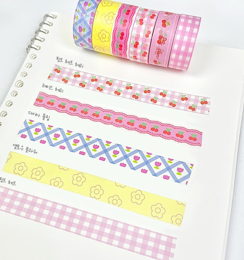 A set of decorative washi tapes with various patterns including cherry prints, gingham, and geometric designs, shown both rolled up and laid out on a white spiral notebook page.