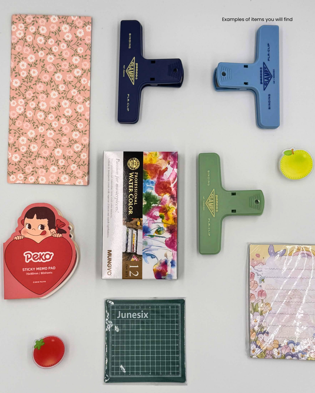 A flat-lay collection of various stationery and craft tools on a light gray background, including a floral sticky pad, blue and green paper clips, a red Peko character sticker, a watercolor paint box, a green cutting mat, and other small accessories.