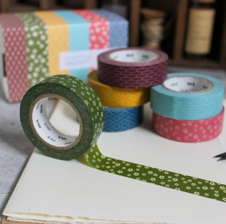 A set of colorful patterned washi tapes in various rolls, with one green patterned roll unspooled in the foreground on a white notebook, set against a blurred background.