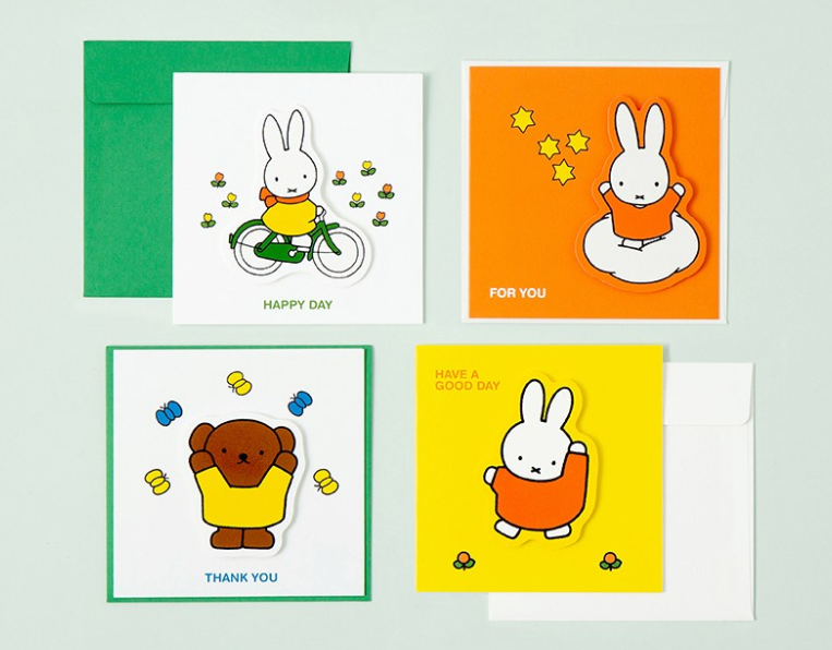 Four Miffy-themed greeting cards with matching envelopes arranged on a pale green background. The cards feature colorful illustrations of Miffy on a bicycle, reading 'HAPPY DAY' and 'FOR YOU', a teddy bear holding a yellow blanket with 'THANK YOU', and Miffy in orange with small clovers and 'HAVE A GOOD DAY'.