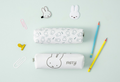 Two white pencil cases with black bunny illustration lineworks of Miffy. They are arranged on a pale mint background with stationery props including paper clips, pencils, and bunny-shaped accessories.