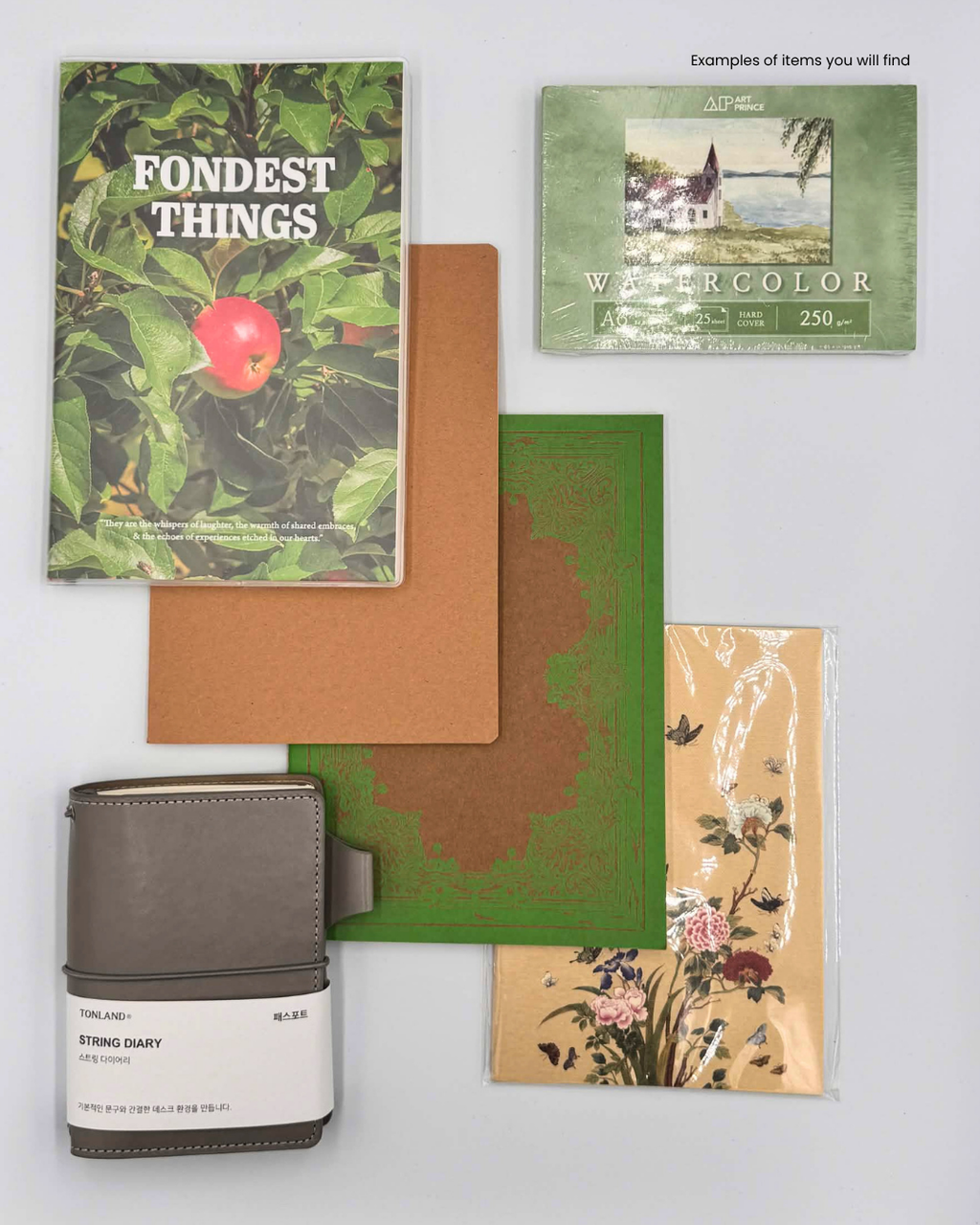 A flat-lay display of various stationery and lifestyle accessories on a light gray background, including a 'Fondest Things' notebook with an apple cover, a watercolor card, a brown textured card, a green card, a beige floral card, and a gray leather-bound diary with a white paper label.