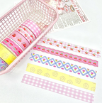 A set of decorative washi tapes with various patterns including cherry motifs, gingham, and geometric designs, displayed inside a pink wire basket and laid out on a white surface next to a vintage-style balloon print.
