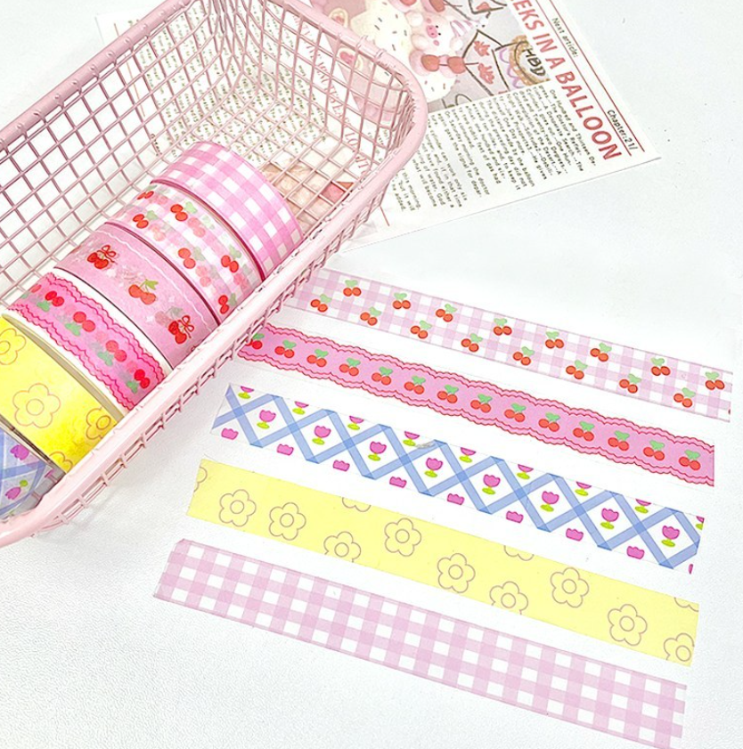 A set of decorative washi tapes with various patterns including cherry motifs, gingham, and geometric designs, displayed inside a pink wire basket and laid out on a white surface next to a vintage-style balloon print.